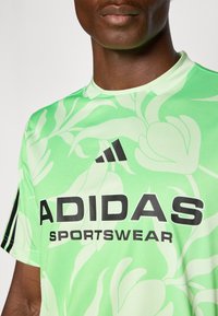 Žalias sportinis marškinėliai su gėlių raštu, juodas "ADIDAS SPORTSWEAR" logotipas, apvaliu kaklu ir juodomis akcentų juostomis ant rankovių.