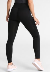 Zwarte high-waisted legging van rekbare stof, met een gladde textuur en een strakke pasvorm. Gecombineerd met lichtroze sportschoenen.