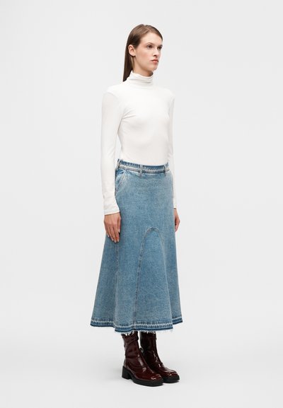 Midi φούστα από τζιν με φαρδιά γραμμή, ακατέργαστο τελείωμα και μπλε χρώμα, συνδυασμένη με εφαρμοστό λευκό ζιβάγκο και γυαλιστερά καφέ μποτάκια στον αστράγαλο.