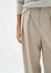 Pantaloni beige su misura con pieghe e una superficie di tessuto liscia. Una mano si appoggia di lato, evidenziando la vestibilità del capo.