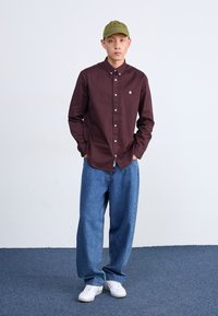 Camisa de botones en color burdeos con botones blancos y logo, combinada con jeans de mezclilla azules holgados y zapatillas blancas. Una gorra verde completa el look.
