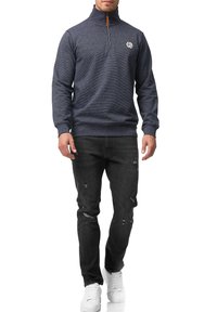 Marineblå ZIP-hoodie med høj krave, udstyret med et lille hvidt logo på brystet, parret med slidte sorte jeans og hvide sneakers.