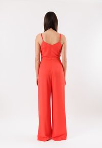 Tuta rossa con top aderente, pantaloni a gamba larga e spalline regolabili. Presenta una chiusura con zip sul retro e una texture di tessuto liscia.