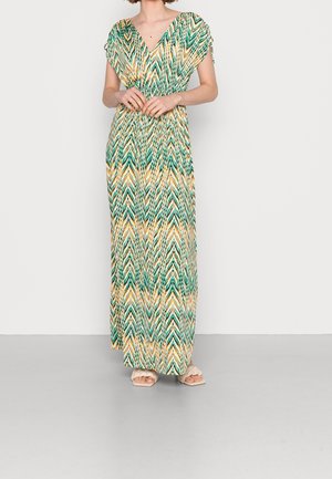Femme portant une robe maxi à motif chevron vert, jaune et blanc, à manches courtes et des sandales blanches, debout, les mains jointes.