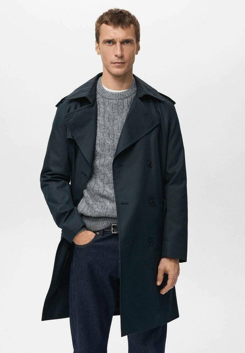 Abrigo gabardina azul marino con diseño de doble botonadura, cuello ancho y detalles de botones, combinado con un suéter gris de punto trenzado y jeans oscuros.