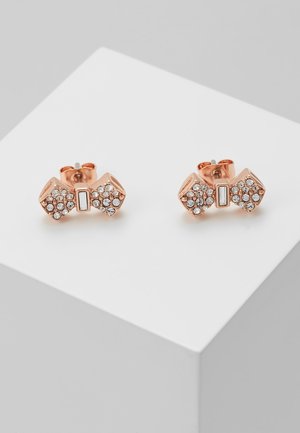 Boucles d'oreilles clous en forme de nœud en or rose ornées de petits cristaux clairs, présentées sur un cube blanc sur un fond gris clair.