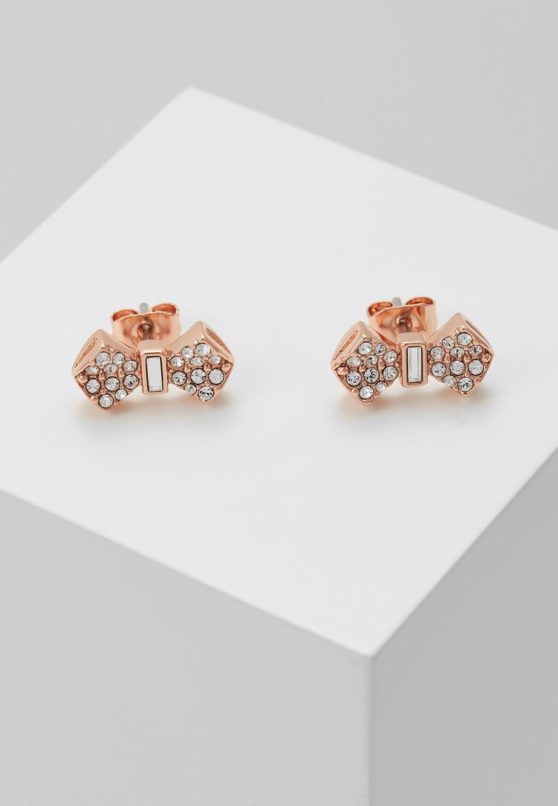 Boucles d'oreilles clous en forme de nœud en or rose ornées de petits cristaux clairs, présentées sur un cube blanc sur un fond gris clair.
