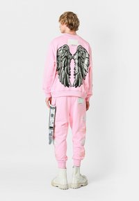 Sudadera rosa con un gran diseño de alas negras en la parte posterior, combinada con pantalones rosa a juego que presentan acentos desgastados y una etiqueta.