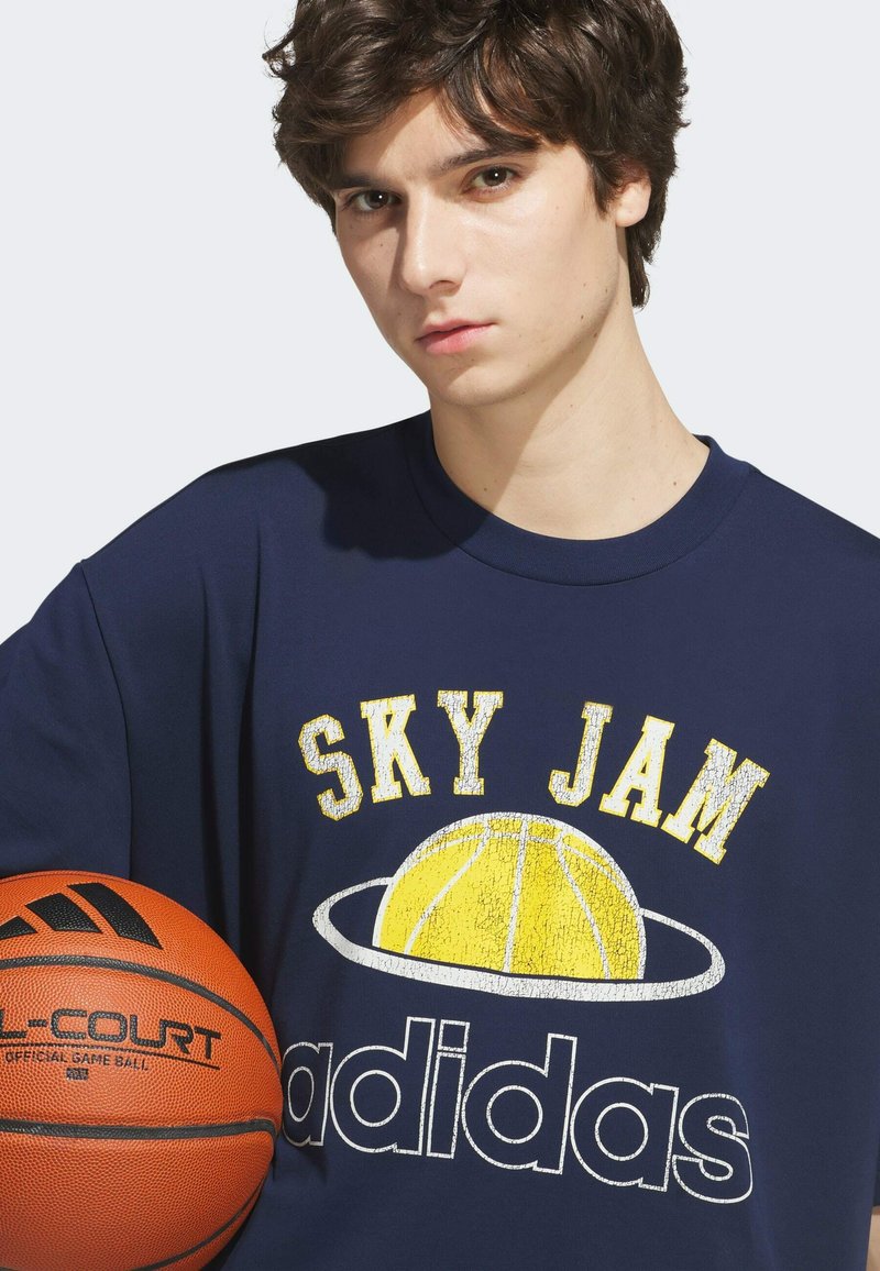 Jeune homme tenant un ballon de basket, portant un t-shirt bleu marine "Sky Jam Adidas" avec un graphisme de basket.