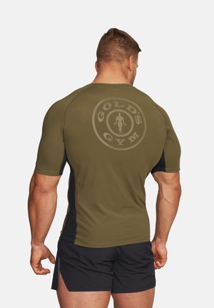 Mężczyzna o muskularnej sylwetce, ubrany w oliwkowozieloną koszulkę Gold's Gym i czarne szorty, widziany od tyłu na jednolitym tle.