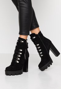Stivaletti neri in suede con tacco spesso, lacci frontali con accenti metallici, zip laterale e suola piatta texturizzata.