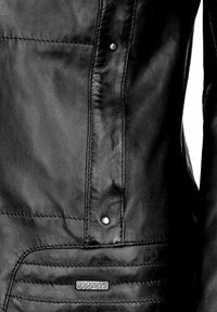 Schwarze Lederjacke mit glatter Textur, ausgestattet mit silbernen Druckknöpfen und einem markierten Metalletikett. Verfügt über genähte Nahtdetails an der Seite.
