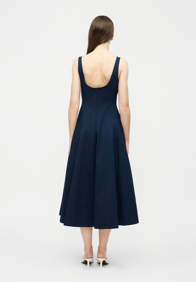 Γυναίκα με μακριά καστανά μαλλιά, που φοράει ένα αμάνικο μεσάτο φόρεμα σε ναυτικό μπλε χρώμα με χαμηλή πλάτη και λευκά ψηλοτάκουνα παπούτσια, στέκεται γυρισμένη προς τα πίσω.