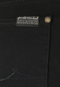 Vaqueros de mezclilla negra con costuras en contraste sutil, una etiqueta en el bolsillo frontal que dice "para toda la humanidad" y un acento dorado en el botón.