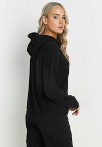 Felpa nera con cappuccio, caratterizzata da una texture a coste, vestibilità ampia e spalle scese. Presenta una tasca a marsupio e una silhouette rilassata.