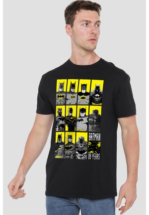 Mann trägt schwarzes T-Shirt mit zwölf verschiedenen Batman-Illustrationen und dem Text "Batman 80 Years" auf der Vorderseite.