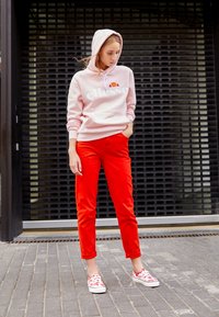 Sweat à capuche rose clair avec logo "ellesse", associé à un pantalon rouge vif. Porte des chaussures à motifs avec une base blanche et des accents rouges.
