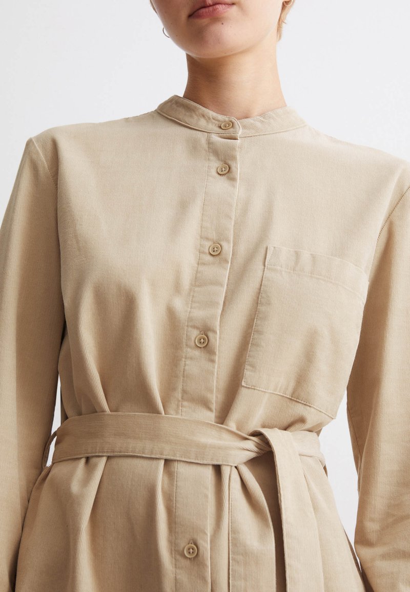 Beiges Hemdkleid mit Mandarin-Kragen, Fronttasche und gebundenem Taillengürtel. Aus strukturiertem Stoff mit einem weichen, lockeren Schnitt.