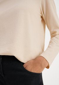 Long-sleeve beige top with subtle vertical stripes, made of soft fabric, paired with black jeans; hand resting in pocket.