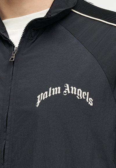 Mustast lukustatavast jopesest lähedalt, millel on valged "palm Angels" tekst gooti kirjas rinnal ja valge torudetail õlal.