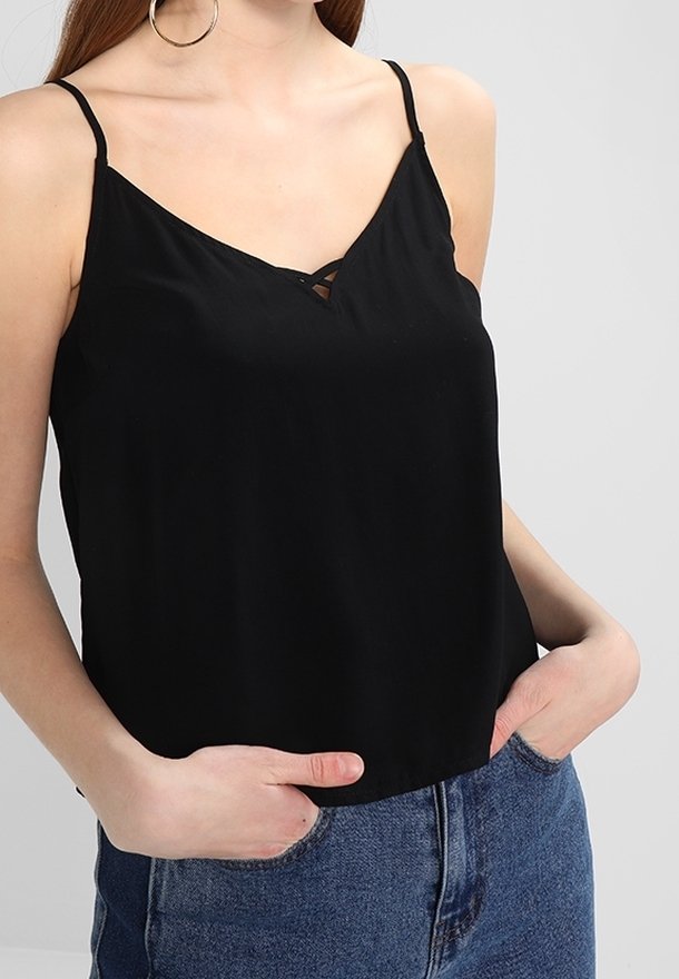 Mujer con top negro sin mangas de escote en V y tirantes finos, y jeans azules de talle alto, con las manos en los bolsillos, sobre un fondo liso.