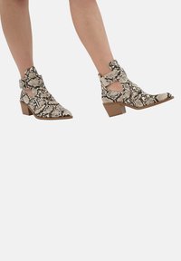 Botas de tobillo de piel de serpiente con tacón cuadrado. Presentan lados recortados y dobles correas. La paleta de colores incluye beige, negro y blanco. Acabado texturizado.