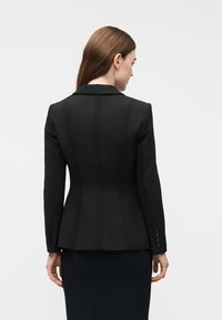 Γυναίκα με ίσια καστανά μαλλιά, φοράει εφαρμοστή μαύρη σακάκι και μαύρη φούστα, γυρισμένη προς τα πίσω μπροστά σε απλό φωτεινό φόντο.