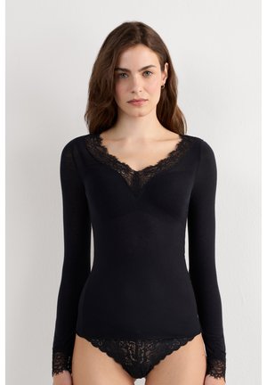 Frau mit langen braunen Haaren trägt schwarzen langärmeligen Top und passende Spitzenunterwäsche vor einfarbigem weißem Hintergrund.