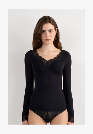 Femme aux longs cheveux bruns porte un haut noir à manches longues et une lingerie en dentelle assortie sur fond blanc uni.