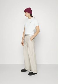 O persoană îmbrăcată cu o cămașă albă cu mânecă scurtă, pantaloni largi bej, pantofi negri și o beretă roșie din tricot, stând în fața unui fundal simplu deschis.