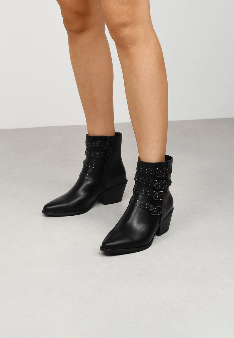 Bottines noires en cuir avec un bout pointu, dotées de trois sangles décoratives à boucle et de clous en argent. Petit talon carré.
