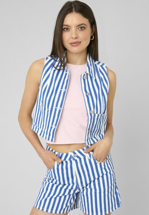 Femme aux longs cheveux bruns portant un gilet sans manches rayé vertical bleu et blanc et un short assorti sur un haut rose clair, mains dans les poches.