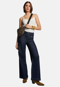 Jeans en denim noir à jambes larges avec poches avant et détails de couture. Associé à un débardeur blanc et un sac à bandoulière à motifs. Bottines à talons marron.