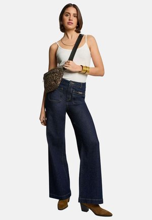 Jeans en denim noir à jambes larges avec poches avant et détails de couture. Associé à un débardeur blanc et un sac à bandoulière à motifs. Bottines à talons marron.