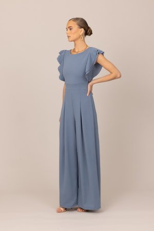 Frau mit zusammengebundenen Haaren, die einen blauen Jumpsuit mit weitem Bein und Volantärmeln sowie goldene Ohrringe trägt, steht mit einer Hand in der Hüfte.