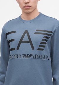 Άνδρας που φοράει μπλε φούτερ Emporio Armani EA7 με μαύρο έντονο λογότυπο και κείμενο στο στήθος, εμφανιζόμενο από τους ώμους μέχρι το μέσο του κορμού.
