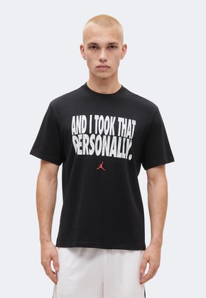 Młody mężczyzna z ogoloną głową, ubrany w czarną koszulkę z białym napisem "AND I TOOK THAT PERSONALLY" i małym czerwonym logo Jordan, stoi nieruchomo.