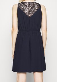 Marineblauwe mouwloze jurk met een aansluitende taille, voorzien van een kant achterontwerp en een soepel materiaal, tot halverwege de dij.
