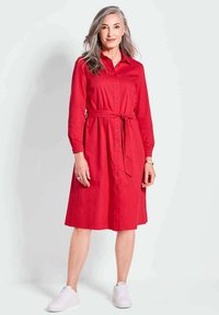 Rotes Hemdblusenkleid mit langen Ärmeln, tailliertem Gürtel und knielangem Saum. Kombiniert mit weißen Sneakern, die eine glatte Stoffoberfläche haben.