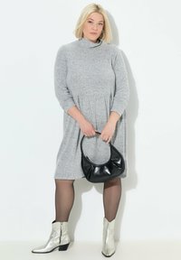 Robe grise, longueur genou, avec un col haut et des manches longues ; associée à un sac à main noir et des bottines argentées. Texture lisse.