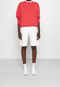 Persona che indossa una felpa rossa a maniche lunghe, pantaloni corti bianchi fino al ginocchio, calze bianche e sneakers bianche, in piedi contro uno sfondo neutro.