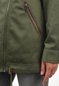 Oliivinvihreä takki, jossa on teksturoitu viimeistely, mukana ruskea vetoketjullinen tasku ja kiristysnauha helmassa. Hihoissa ja helmassa näkyvä ompelu.