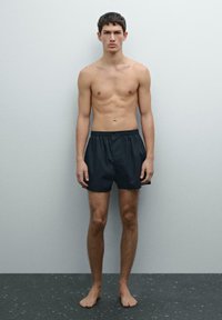 Shorts de bain noirs en matériau léger, dotés d'une taille élastique, de poches latérales et d'une coupe décontractée, présentés sur un modèle masculin devant un fond uni.