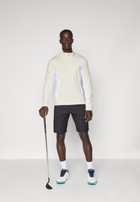 Svetlo béžová golfová košeľa s dlhým rukávom a bielymi bočnými panelmi, v kombinácii s čiernymi šortkami a bielymi golfovými topánkami s námorníckymi akcentmi. Drží golfovú palicu.