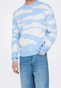 Lichtblauwe trui met witte wolkenpatronen, geribde halslijn en manchetten, gemaakt van gebreid materiaal; gedragen met lichtblauwe jeans.