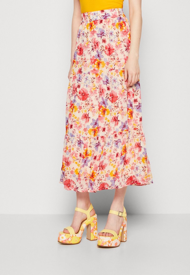 Femme portant une jupe midi rose à fleurs et des talons compensés jaunes à fleurs avec brides à la cheville, debout devant un fond uni.