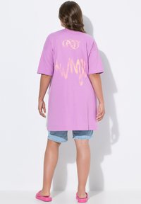 Lila oversized T-shirt met korte mouwen heeft een gestileerde oranje graphic op de achterkant. Gepaarde met blauwe denim shorts en roze sandaaltjes.