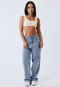 Lichtbeige crop top met logo. Blauwe denim wijde broek met witte stiksels en grote patchdetails. Model staat tegen een effen achtergrond.
