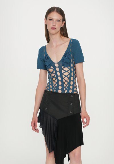 Haut en tricot bleu avec des motifs découpés et des nœuds, associé à une jupe asymétrique noire avec des boutons et des textures superposées.