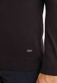 Maglione nero a maglia con bordo e polsini a costine, con un'etichetta in tessuto che riporta la dicitura "DEDICATO ALLA PERFEZIONE DEL DENIM" sul lato inferiore.